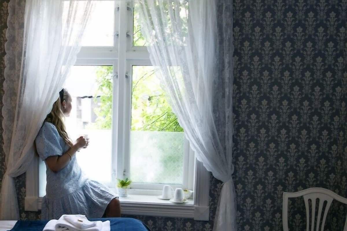 Habitació amb llit, una cadira, finestra amb cortines i vista a vegetació a l'exterior.