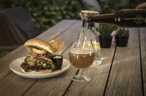 Una comida con una hamburguesa y papas fritas sobre una mesa de madera, una jarra de cerveza y una botella siendo servida.