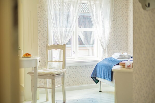 A room with a bed, a chair, a small table, and a window with sheer curtains.