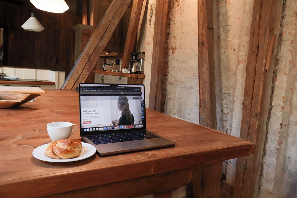 Una laptop sobre una mesa de madera con una taza y un pastel, en una habitación rústica con vigas a la vista y pared de ladrillo.