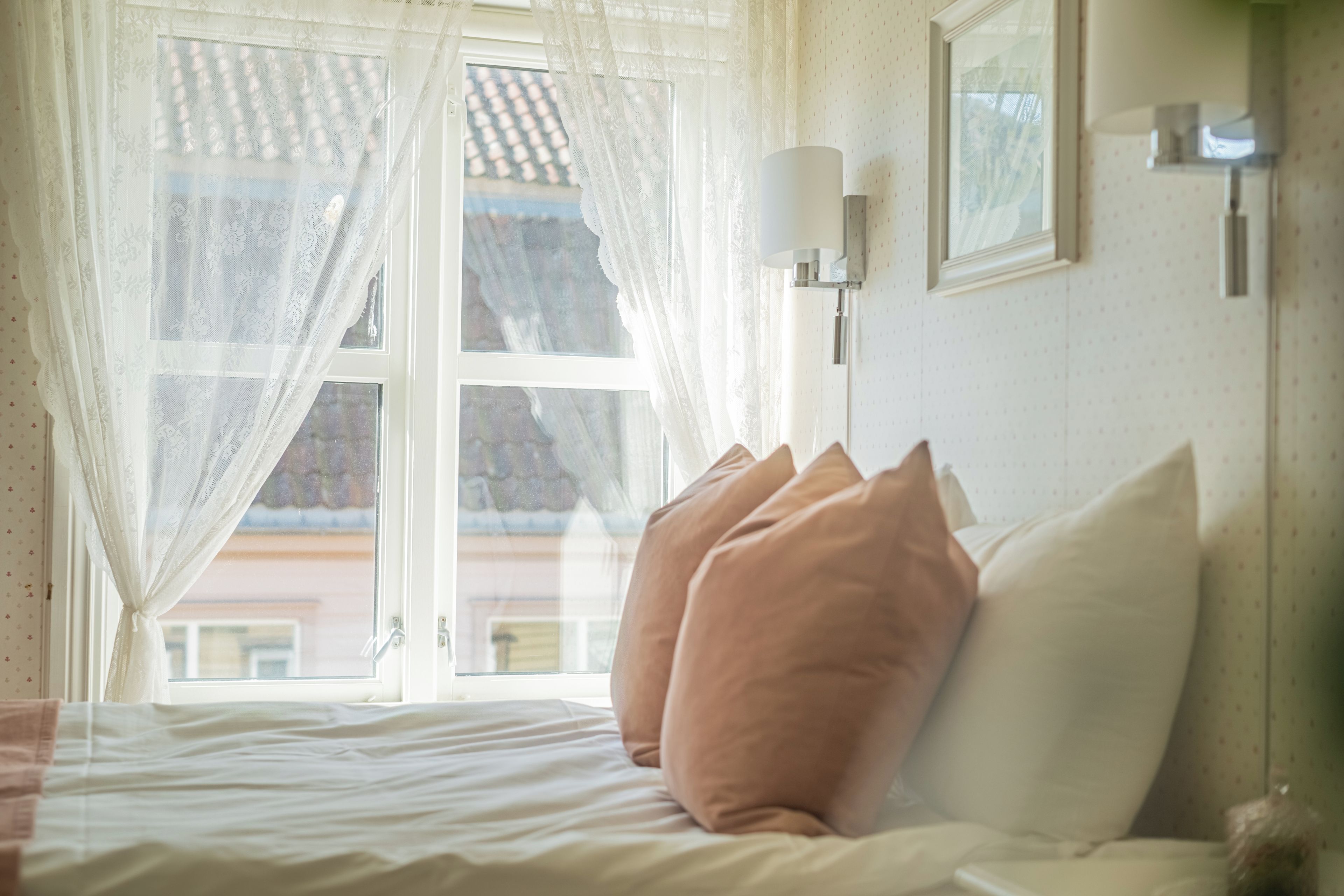 A bedroom with a bed, pillows, a window with lace curtains, and a wall-mounted lamp.