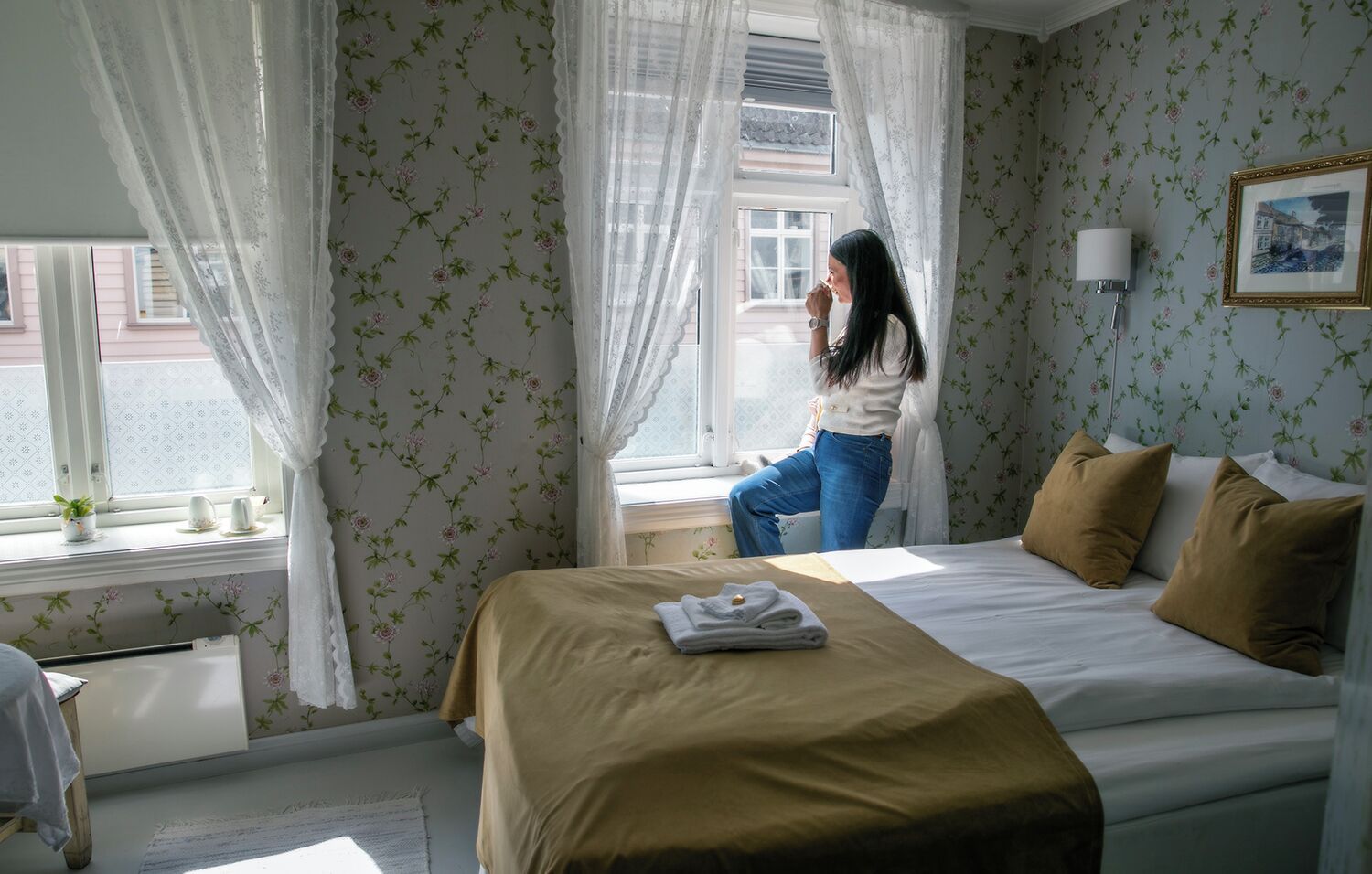 A hotel room with a bed, a window with curtains, a bedside table, and a wall with floral wallpaper.