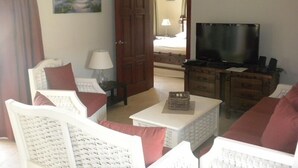 A living room with a television, white wicker furniture, a painting on the wall, and a door leading to another room. - Villa 604 RCL (Sosúa)