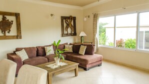 A living room with a brown sofa, a wooden coffee table with a plant, and a large window with curtains. - Well Furnished w Private Pool, BBQ & Palapa Bar (Sosúa)