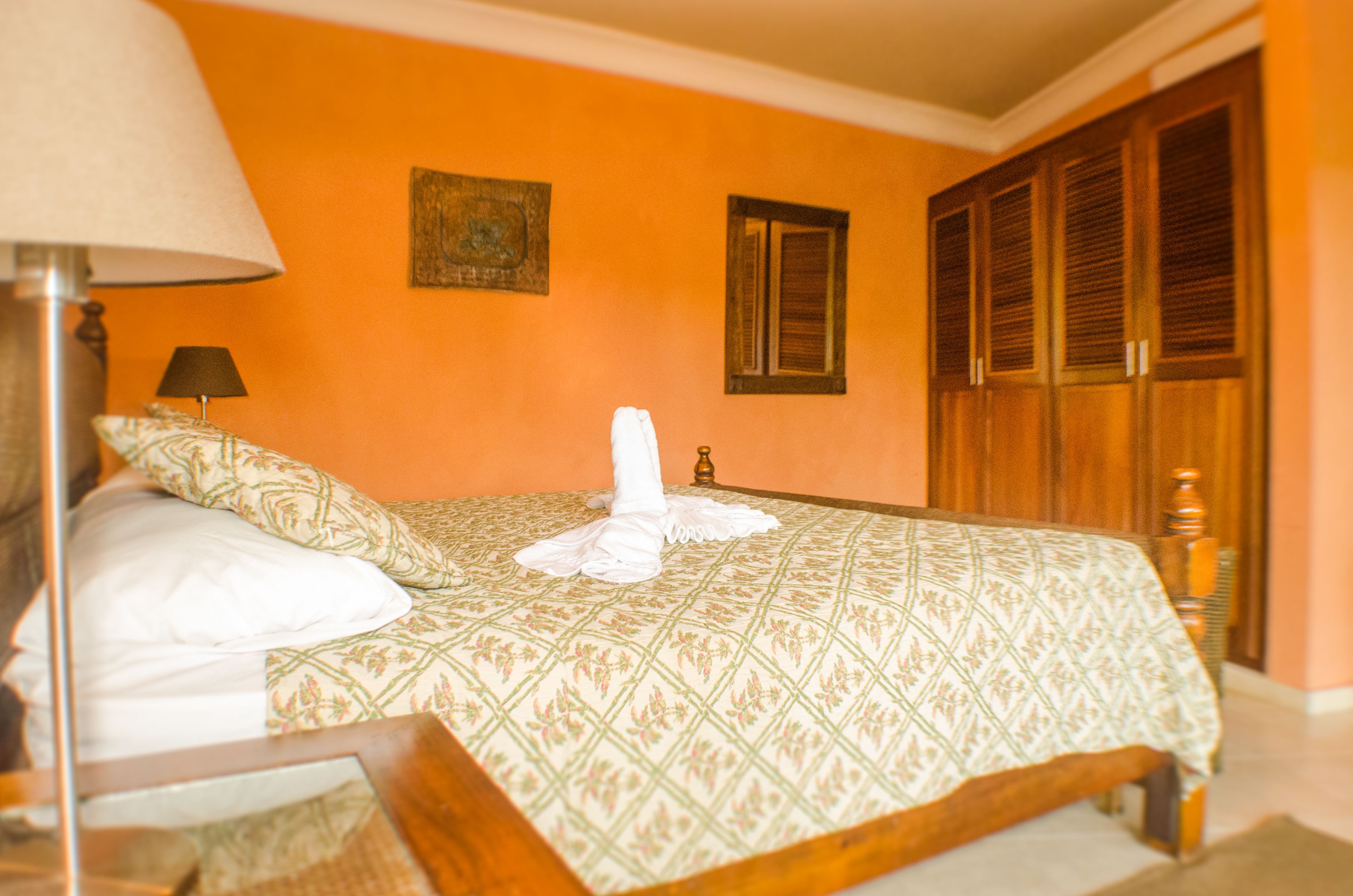 Un lit bien fait, recouvert d’une couvre-lit à motifs, une lampe de chevet et une armoire en bois dans une pièce aux murs orange.