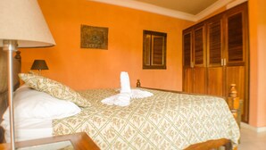 A neatly made bed with a patterned bedspread, a bedside lamp, and a wooden wardrobe in a room with orange walls. - Well Furnished w Private Pool, BBQ & Palapa Bar (Sosúa)