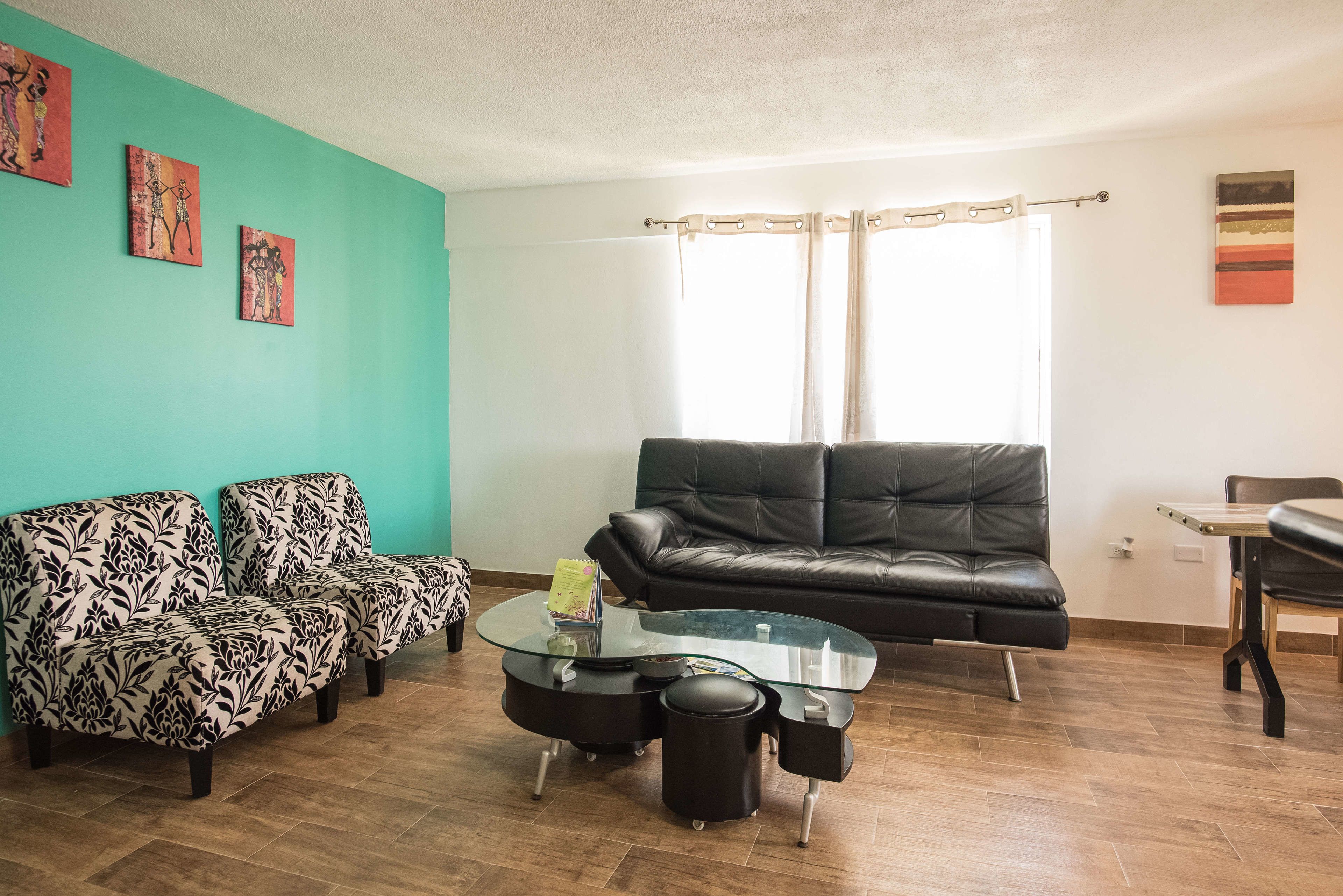 Un salon avec un mur de couleur turquoise, deux canapés à motifs, un canapé en cuir noir, une table basse en verre et une table à manger avec des chaises.