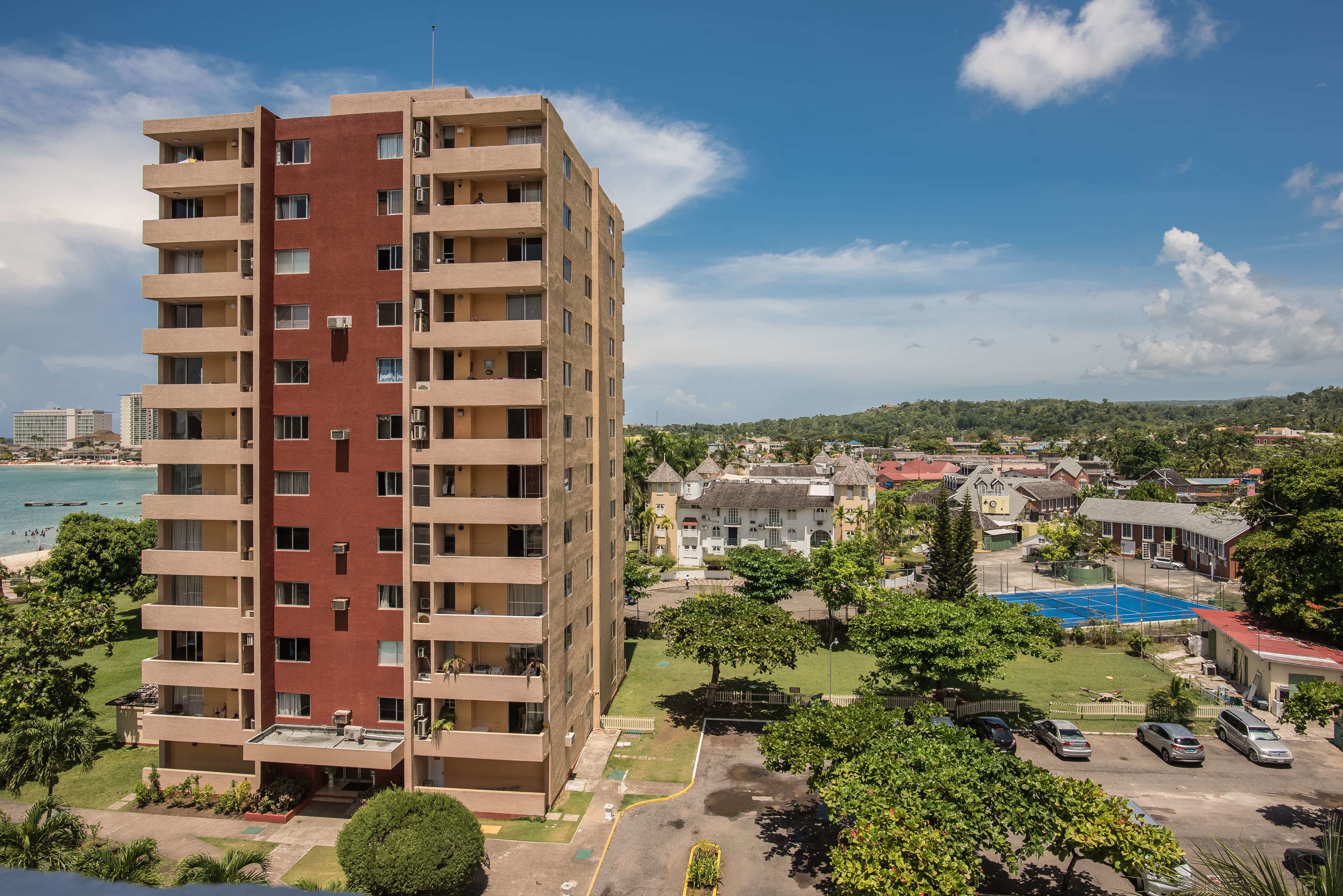 A multi-story building with balconies, a parking area, and a view of a beach and town in the background.