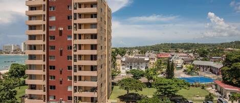 A multi-story building with balconies, a parking area, and a view of a beach and town in the background.