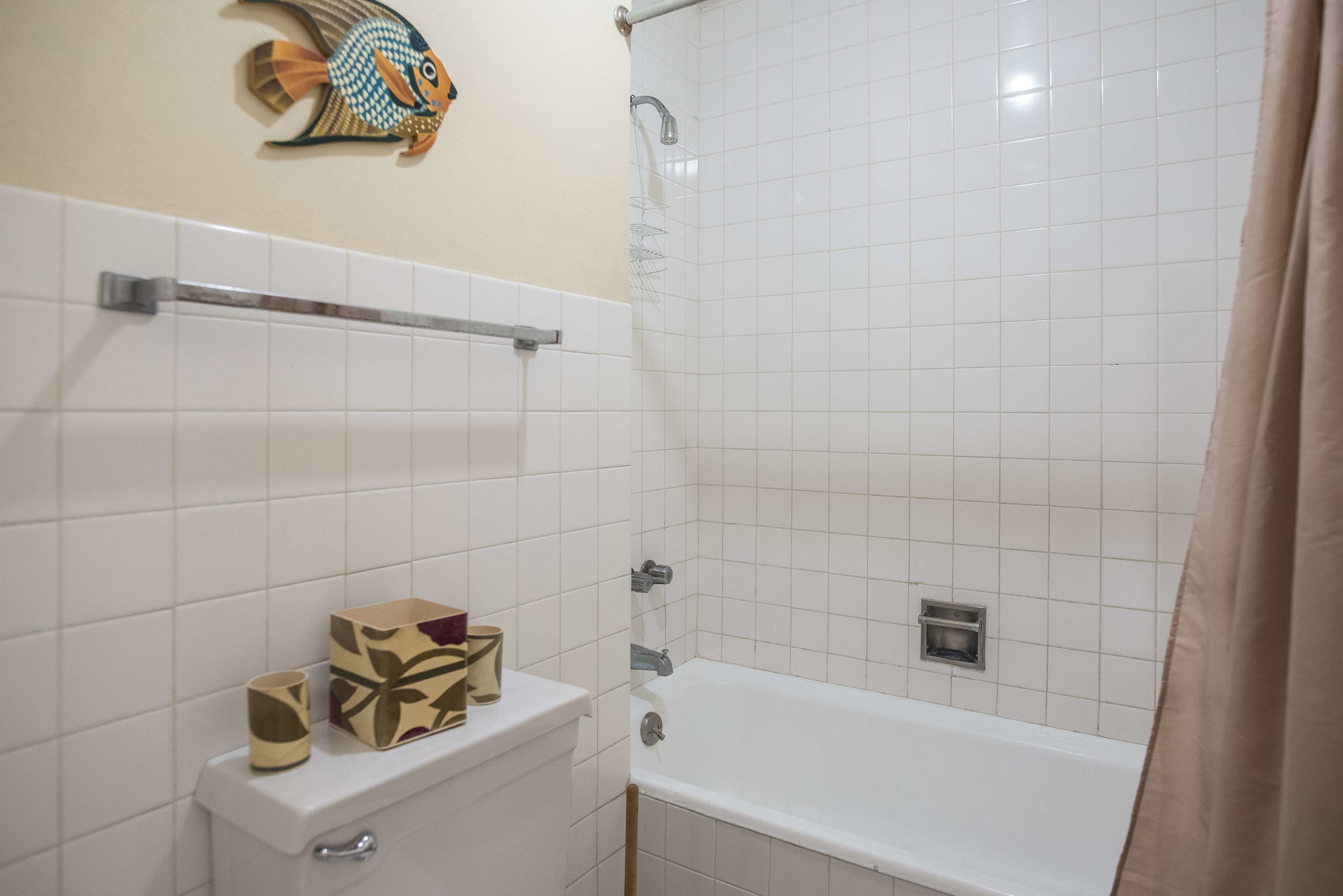 Un baño con paredes de azulejos blancos, una bañera, un inodoro, un perchero y una obra de arte de peces decorativos en la pared.