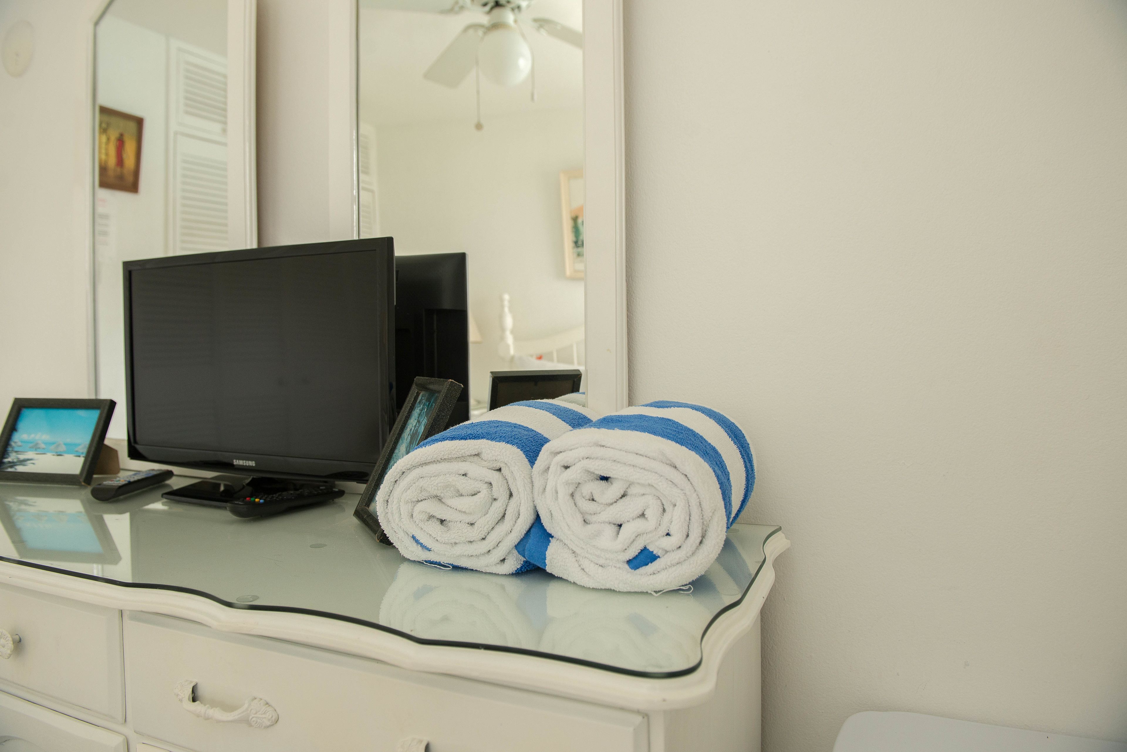 A hotel room with a glass-top desk, a flat-screen TV, a smartphone, and rolled towels.