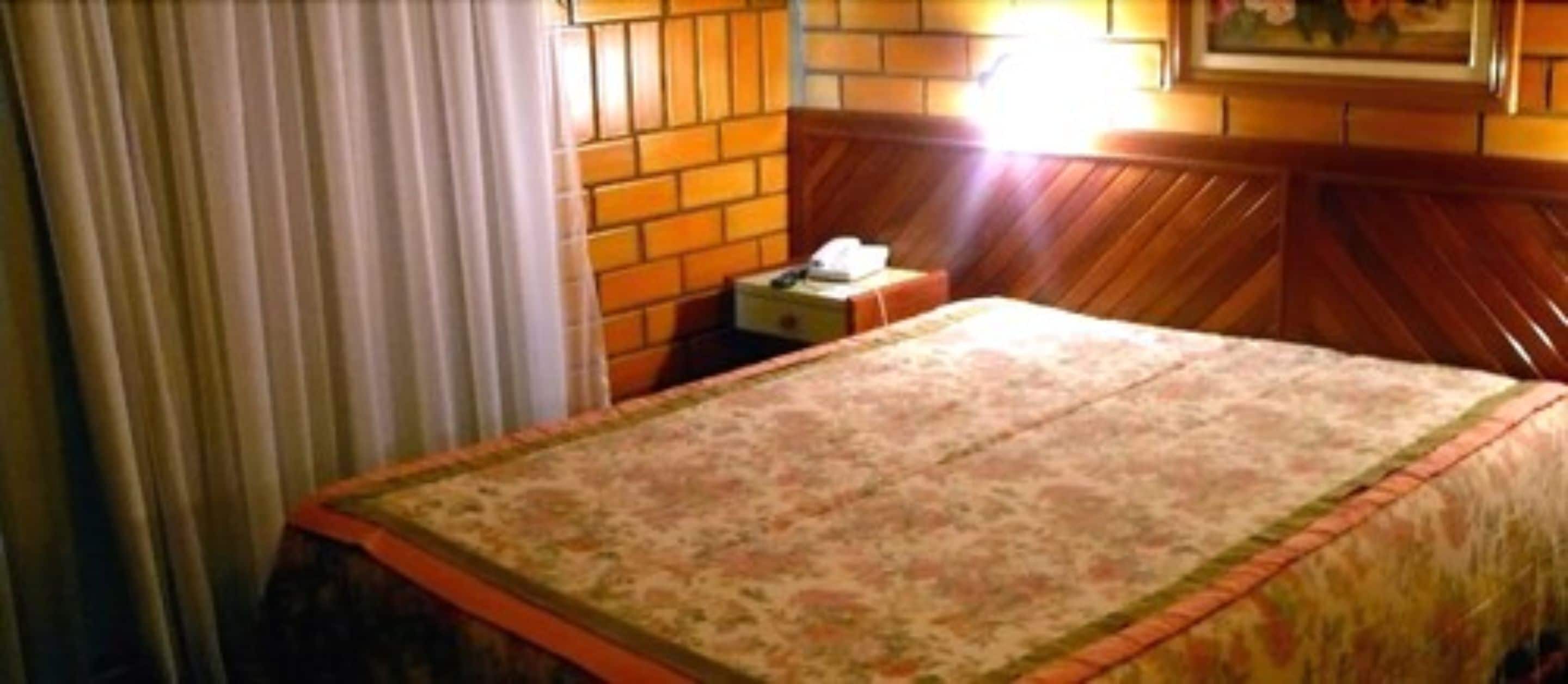 Una habitación de hotel con cama, mesita de noche, lámpara y ventana con cortinas.
