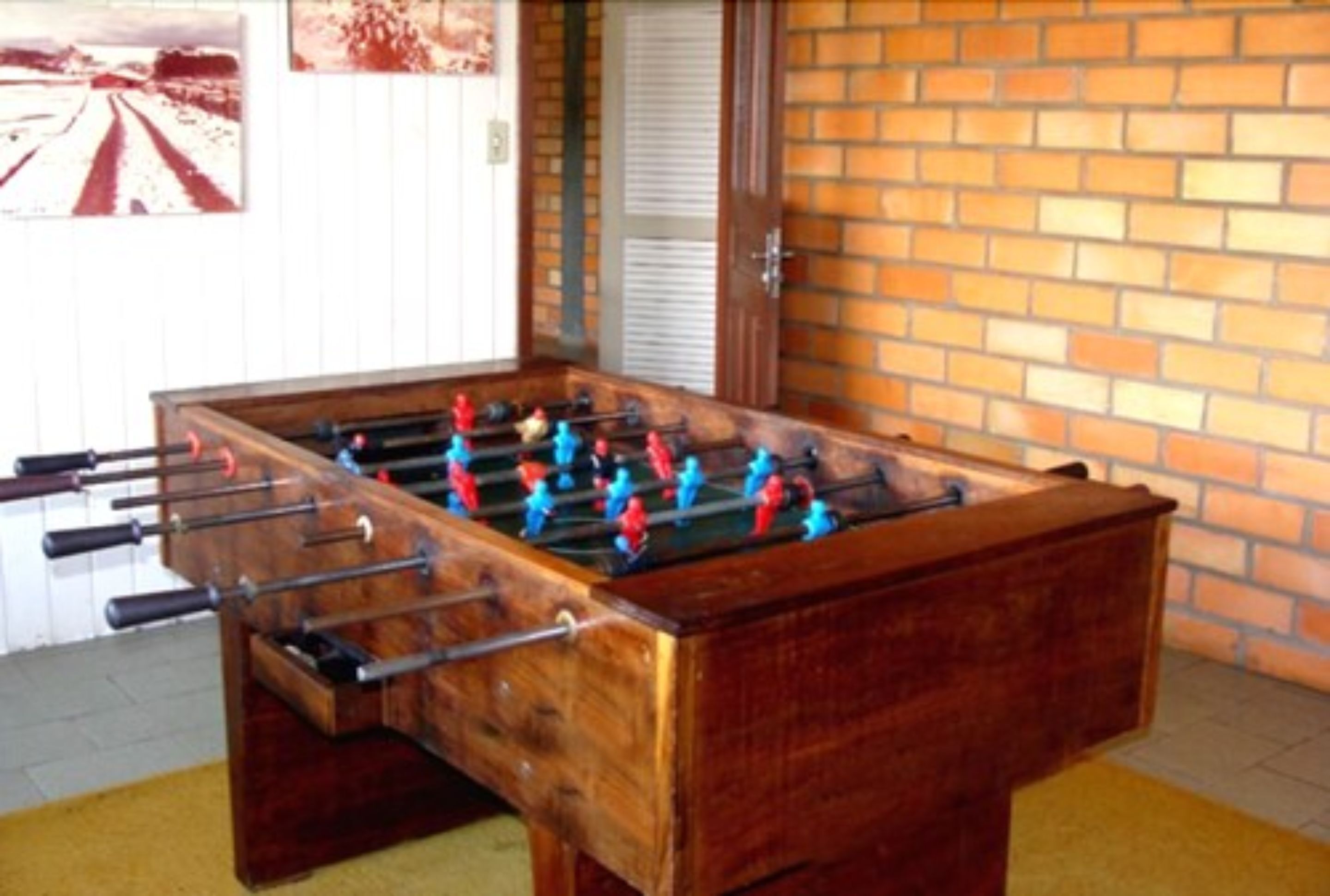 Une table de baby-foot en bois, avec des figurines rouges et bleues, placée devant un mur de briques et une porte blanche.