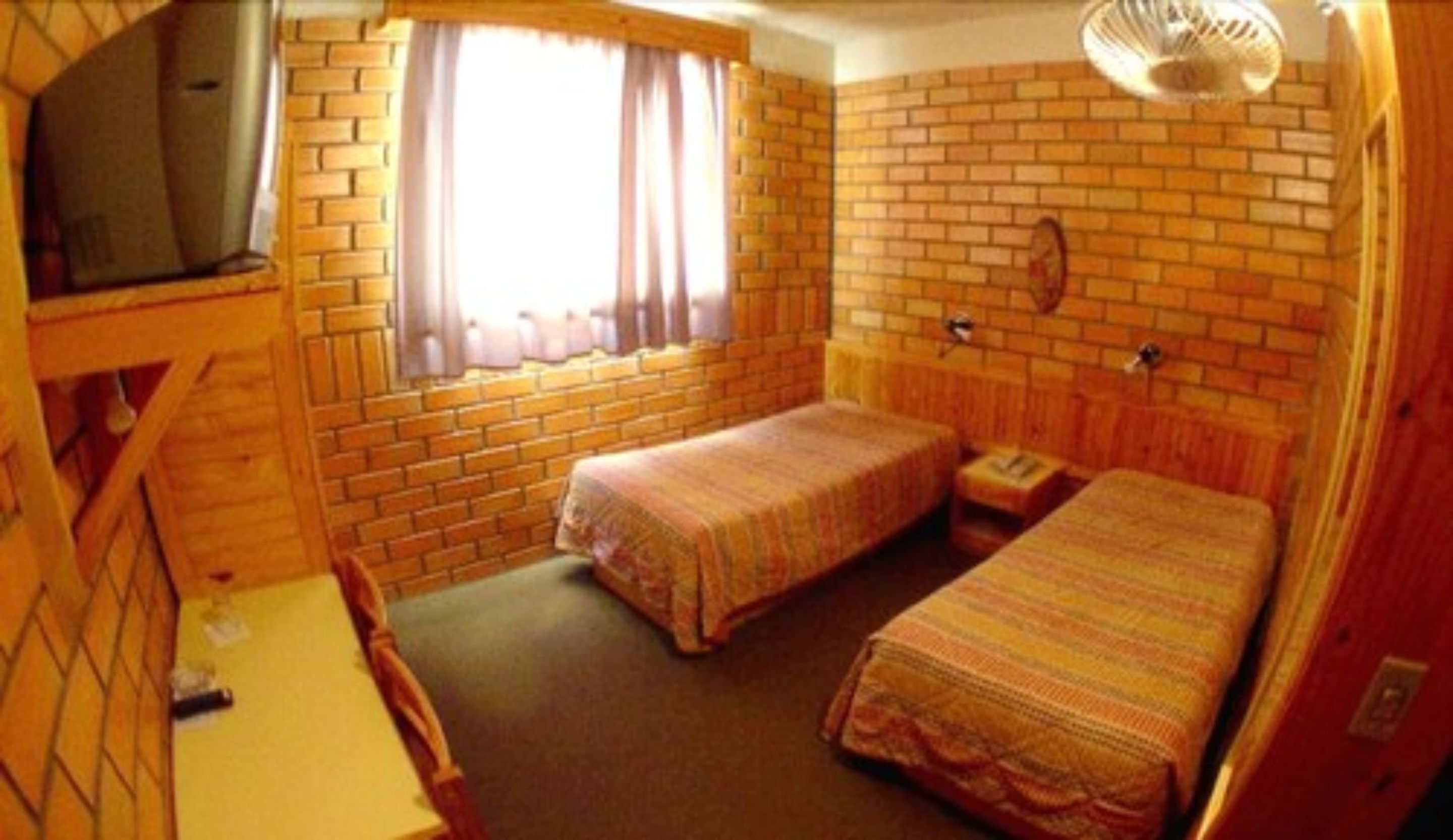 A room with two single beds, a wooden desk, a chair, a television, and a window with curtains.