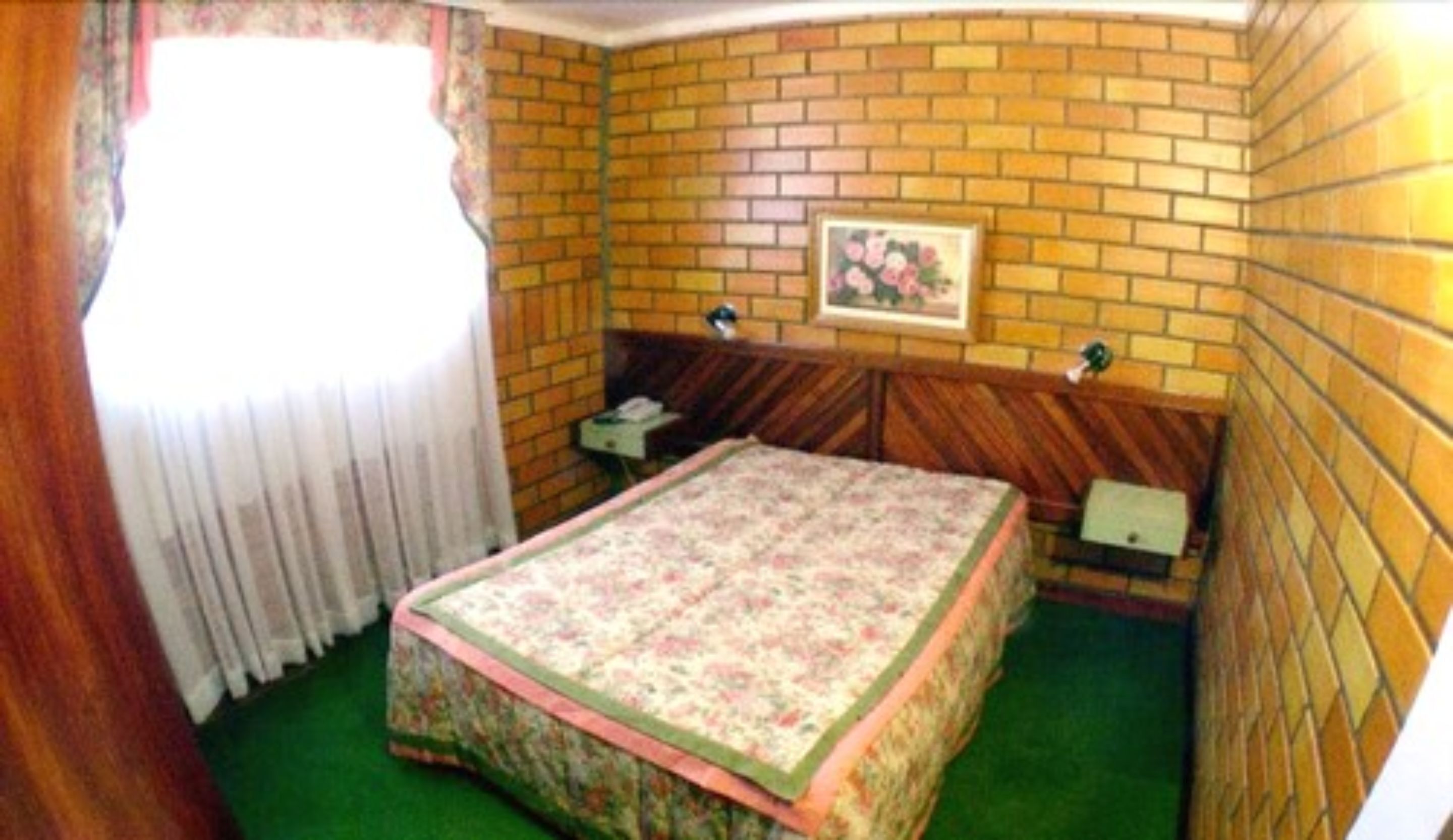 A small, cozy room with a brick wall, a single bed with a floral bedspread, a small table, and a window with curtains.