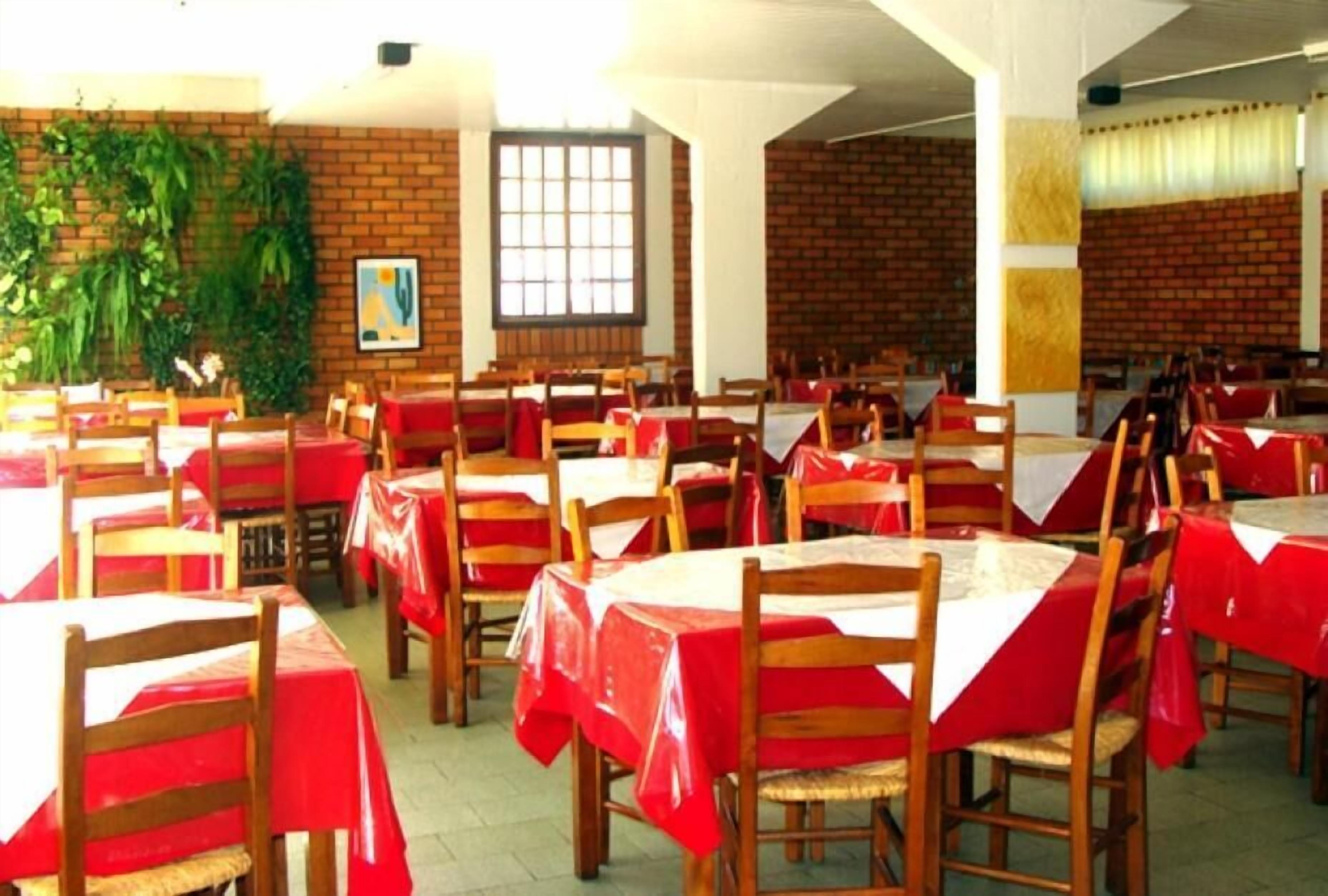 Un comedor con mesas cubiertas de manteles rojos y sillas de madera, paredes de ladrillo y un ventanal grande.