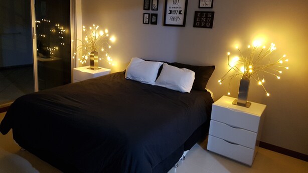 Une chambre à coucher avec une couvre-lit noire, des oreillers blancs et une table de chevet sur laquelle se trouve un vase décoratif avec des lumières. Des tableaux encadrés ornent le mur.