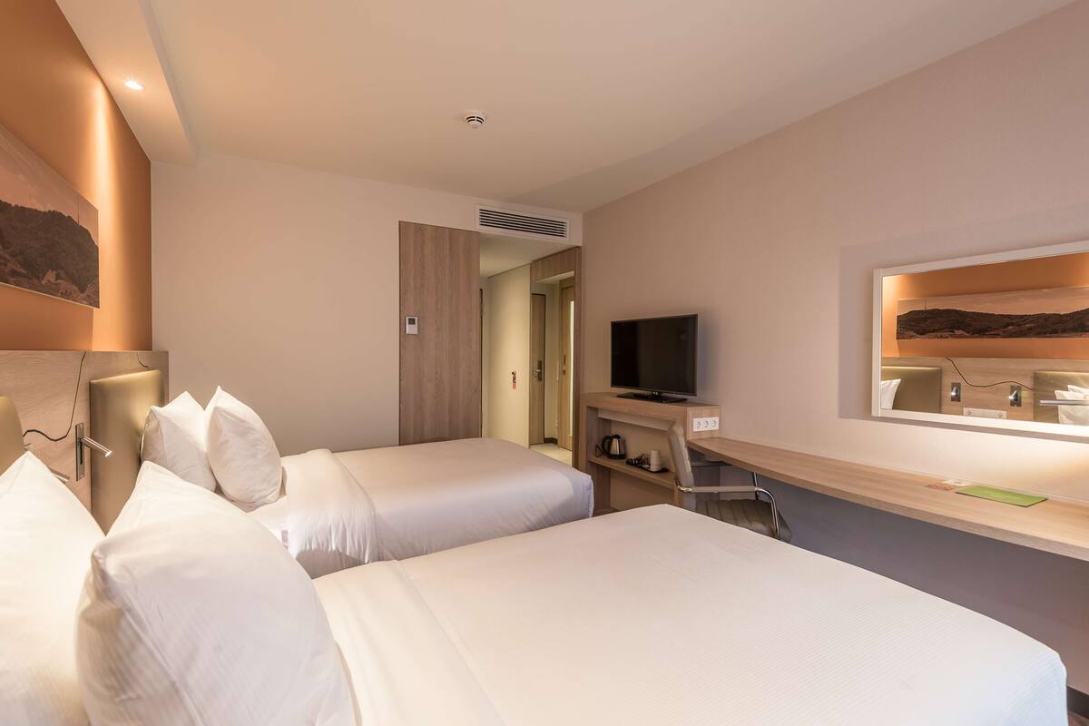 Une chambre d’hôtel avec deux lits simples, une télévision, un bureau avec un miroir et un tableau au mur.