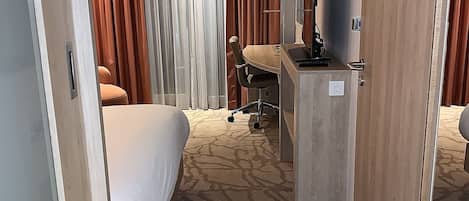 A hotel room with a bed, a desk with a chair, a television, and a window with curtains.