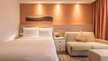 A modern hotel room with a large bed, a sofa, a nightstand, and a wall-mounted artwork.