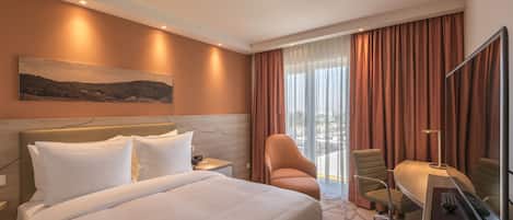 A hotel room with a large bed, a desk with a computer, a chair, and a view of the outdoors through a window with curtains.