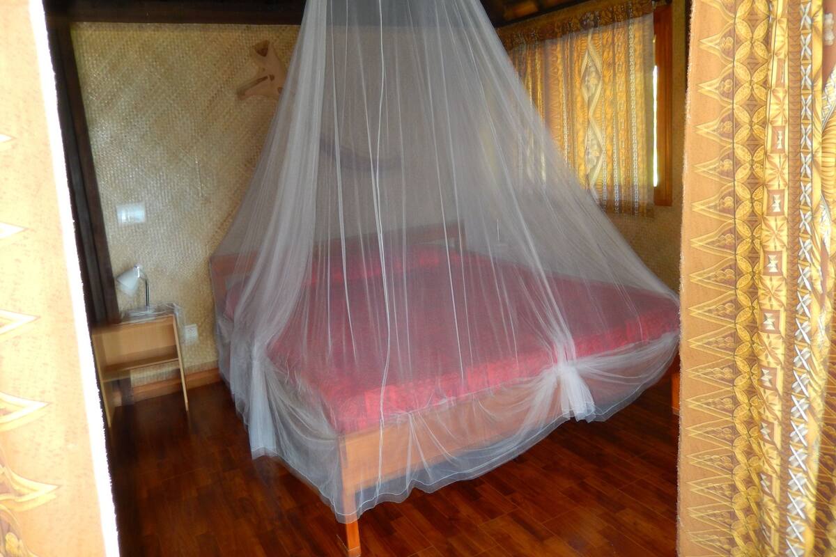 A room with a bed under a mosquito net, wooden flooring, and patterned curtains.