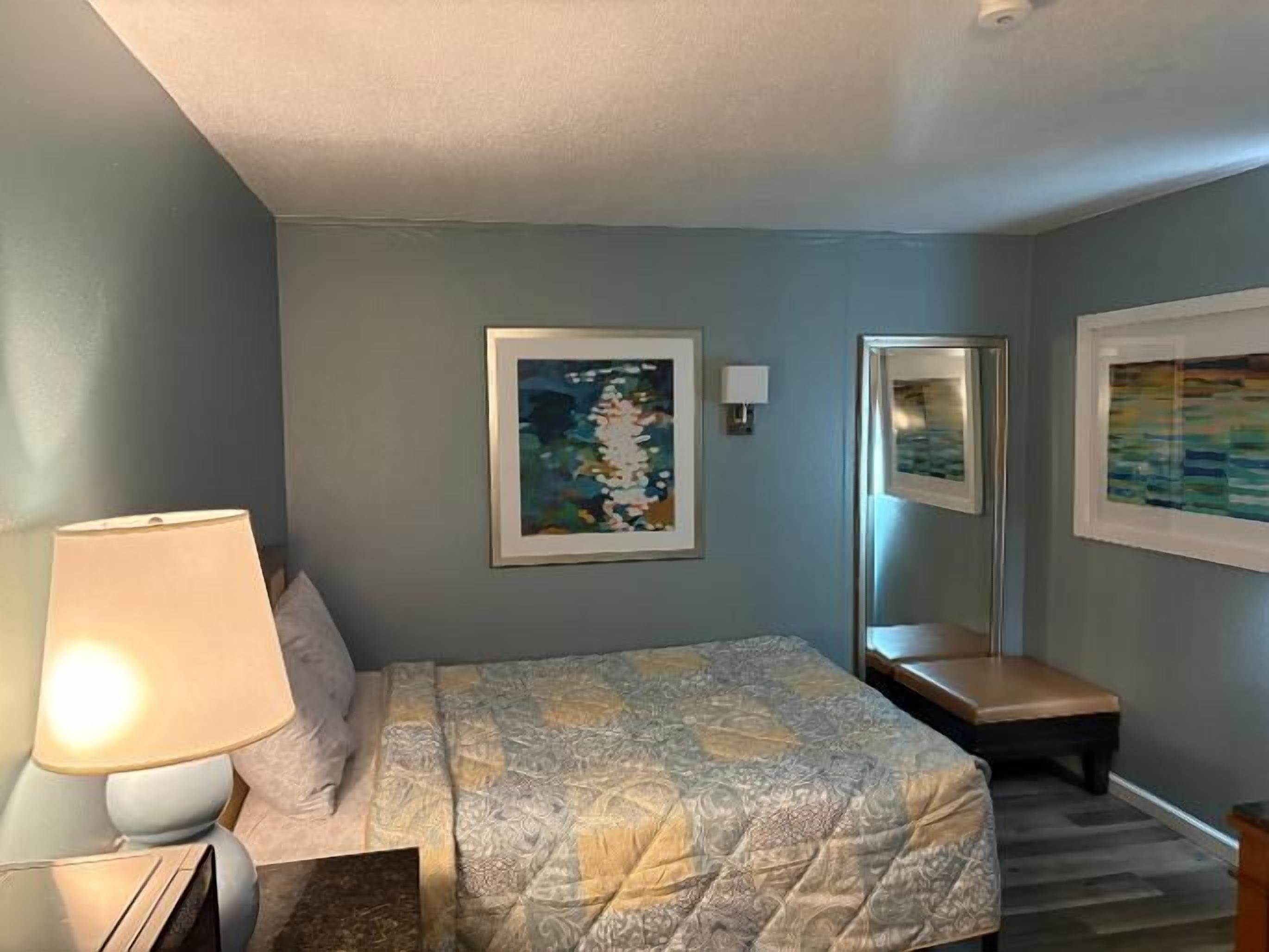 A hotel room with a bed, a bedside lamp, a chair, and two framed artworks on the wall.
