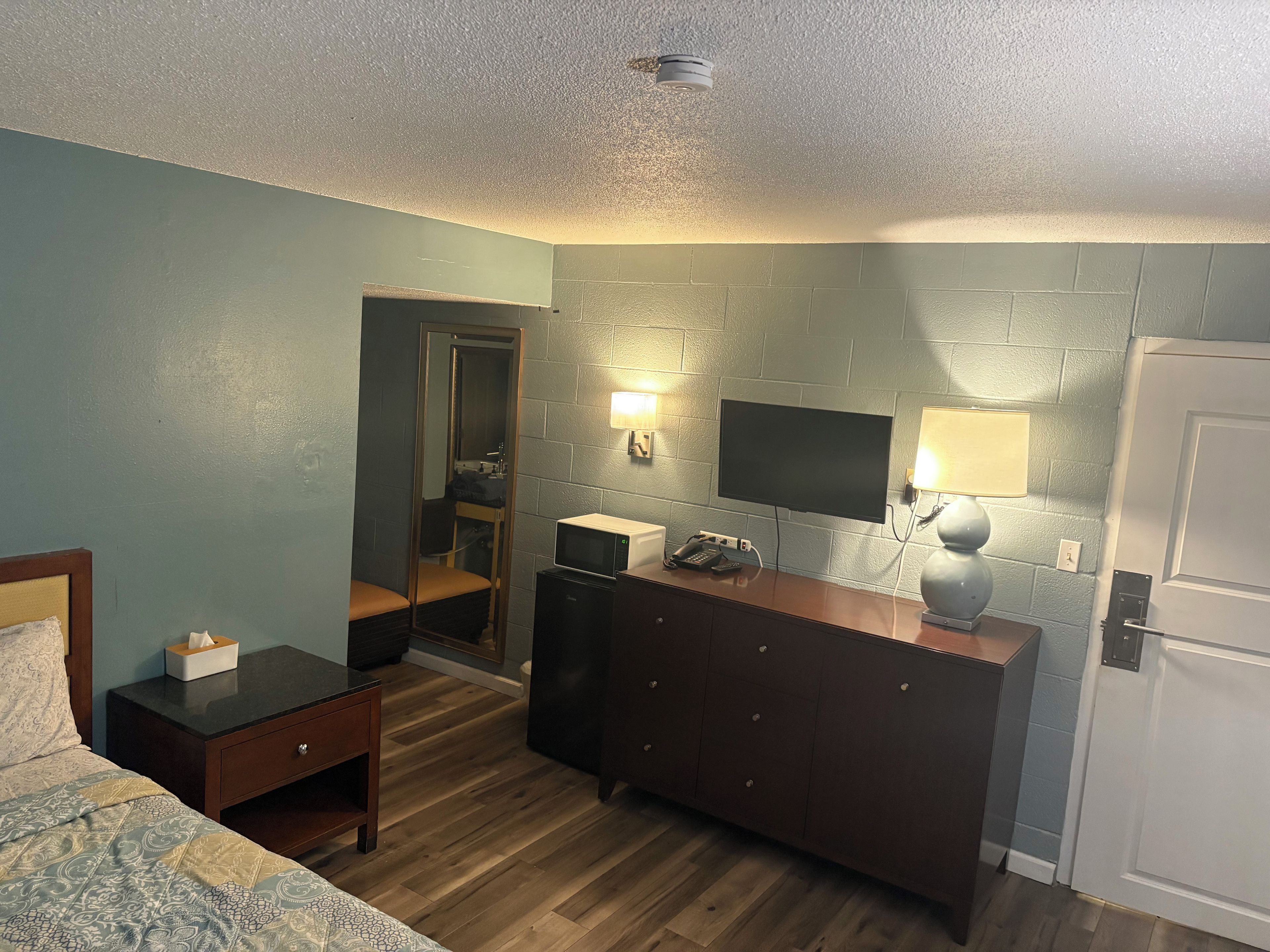Una habitación de hotel con cama, mesita de noche, cómoda, espejo, televisor y microondas.