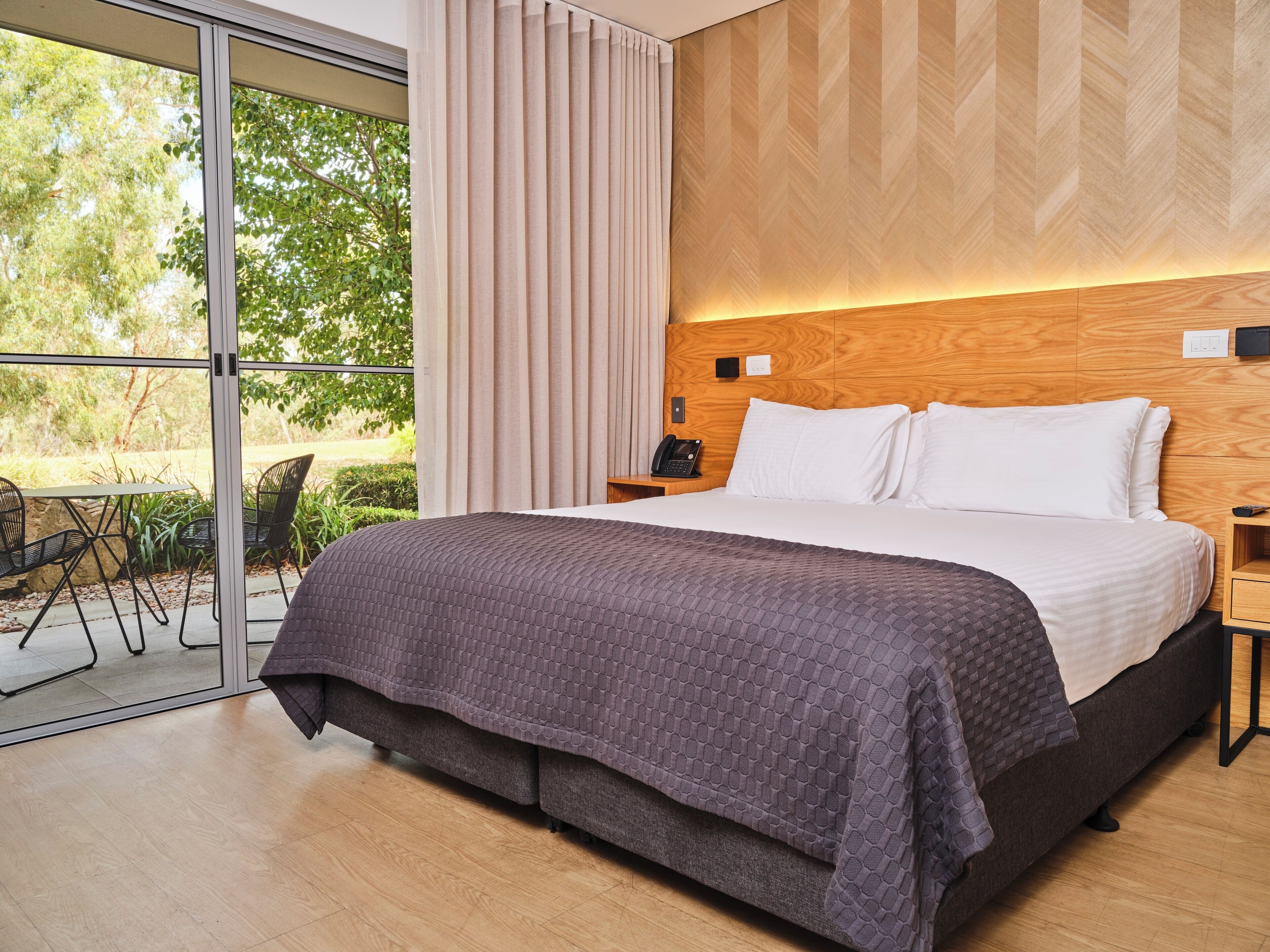 A modern hotel room with a large bed, a wooden headboard, a glass door leading to an outdoor area, and a small table with a phone.