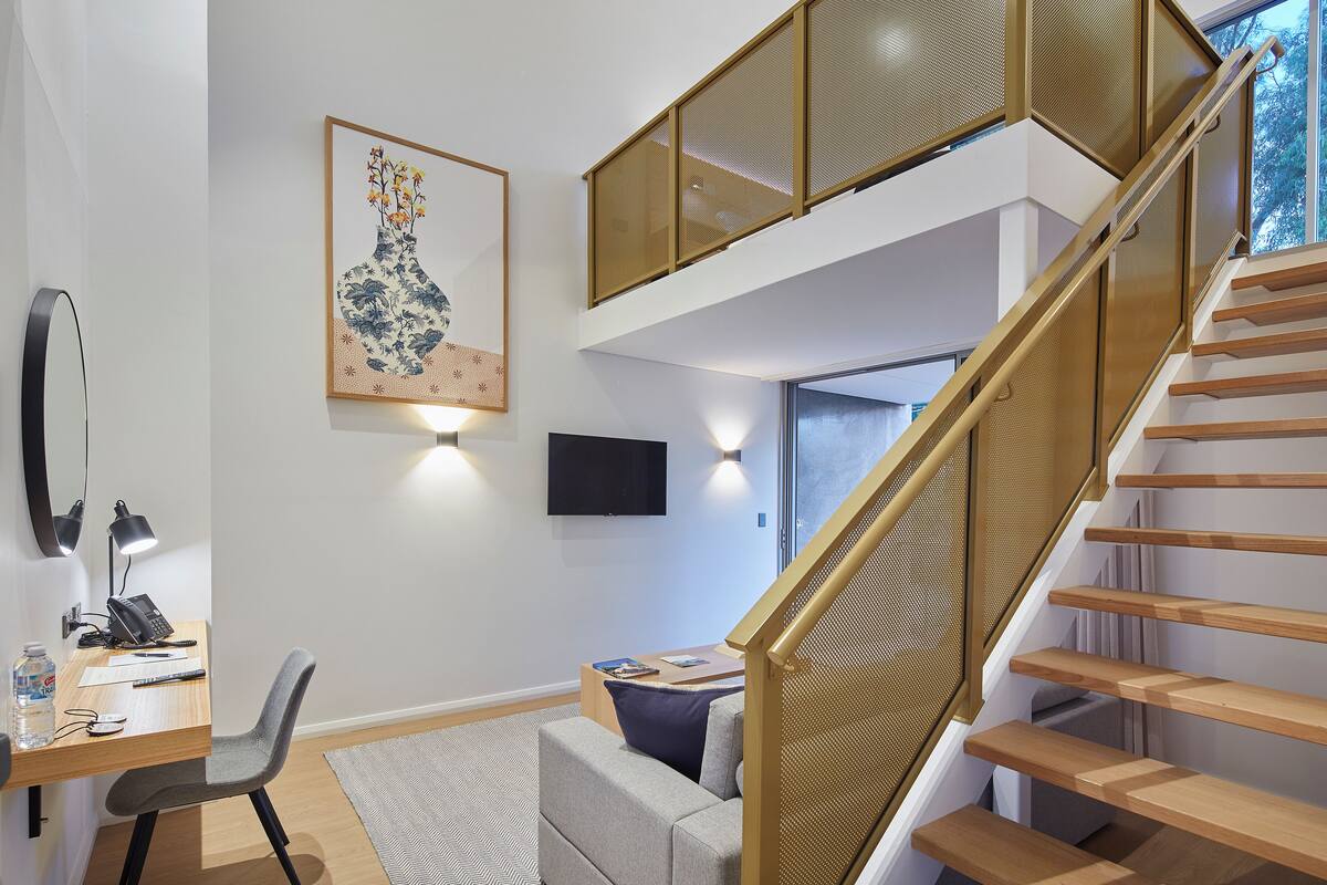 Une pièce moderne et bien éclairée, équipée d’un bureau, d’une chaise et d’un canapé. Un escalier avec une rampe en bois mène à l’étage supérieur.