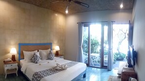 A bedroom with a large bed, two bedside tables with lamps, a TV on the wall, and a view of the outdoors through a sliding door. - Jukung Guest House (Denpasar)