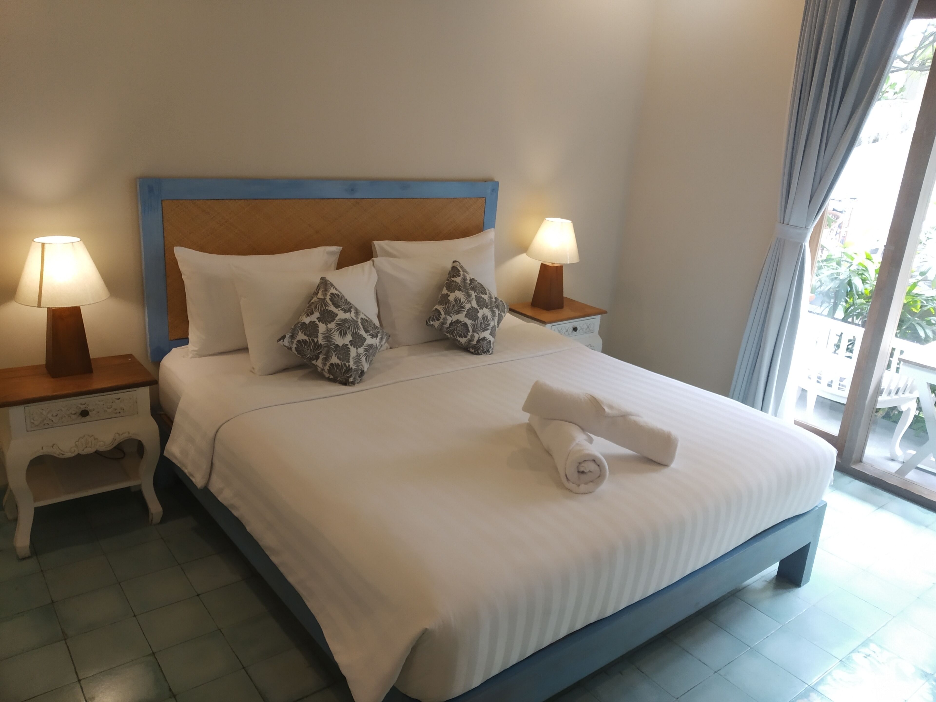Una cama bien hecha con sábanas blancas y almohadas decorativas, dos lámparas de noche y vistas al exterior a través de un ventanal grande.