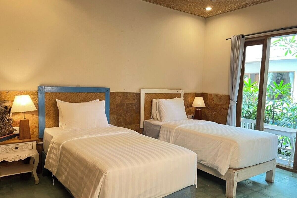 A hotel room with two beds, a nightstand with a lamp, and a view of greenery through the window.