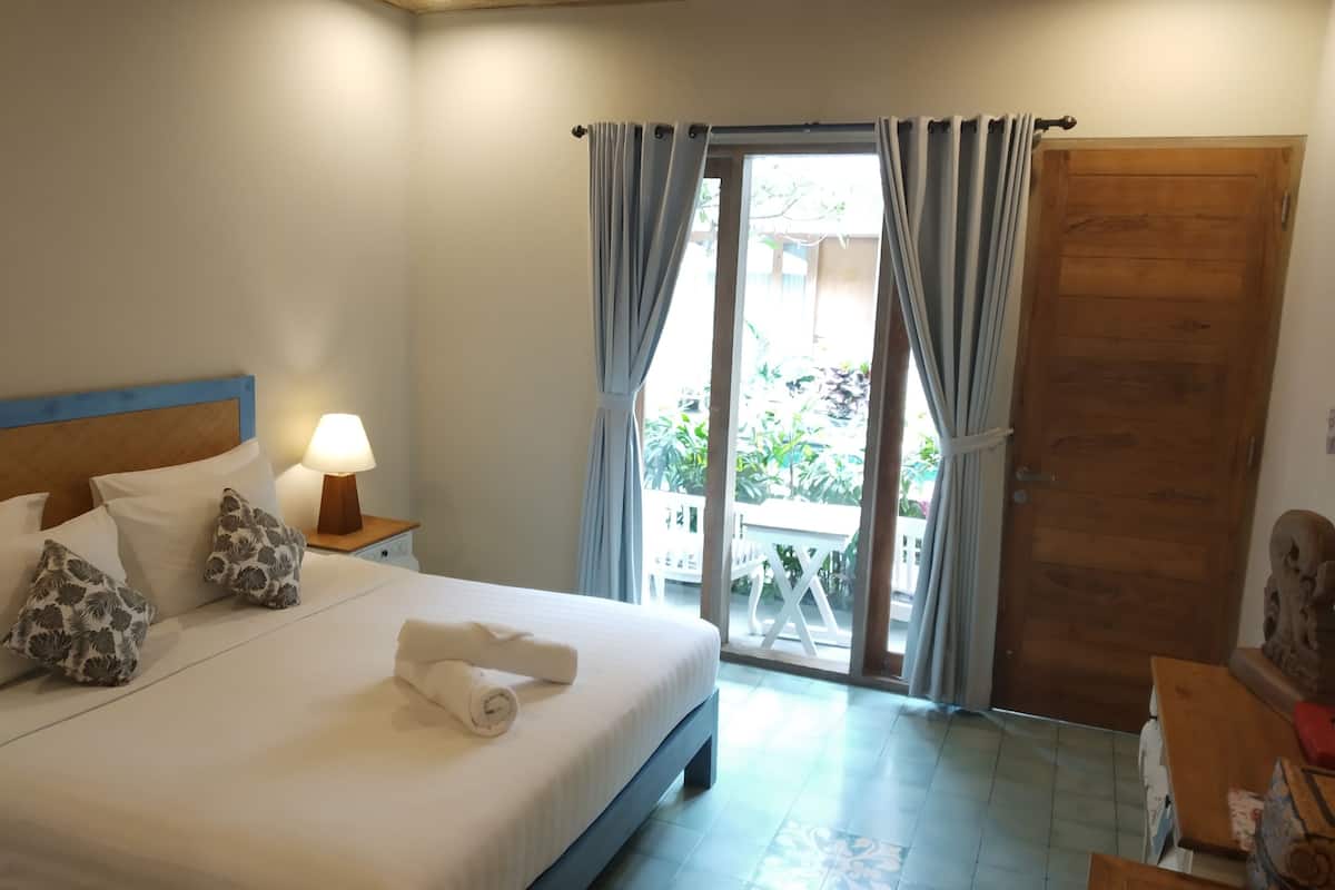 A hotel room with a bed, a nightstand with a lamp, a wooden door, and a view of the outside.