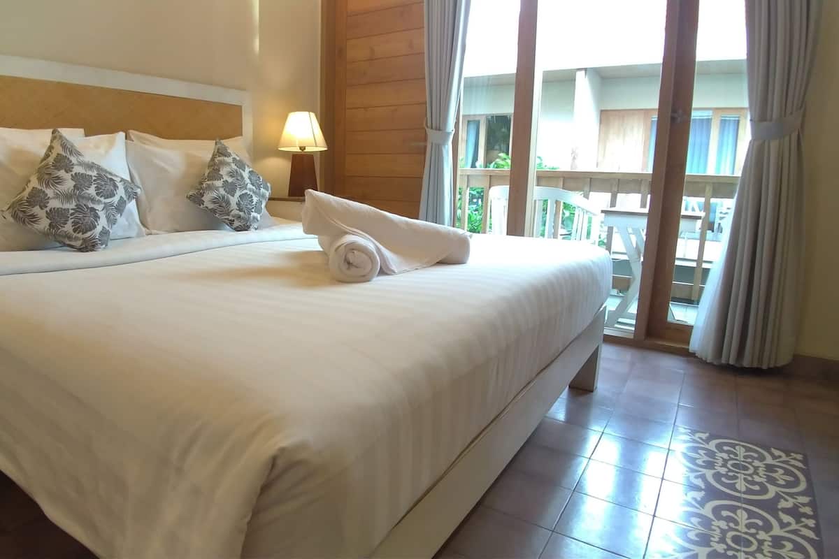 A hotel room with a large bed, a wooden headboard, a nightstand with a lamp, and a view of the outdoors through a sliding door.
