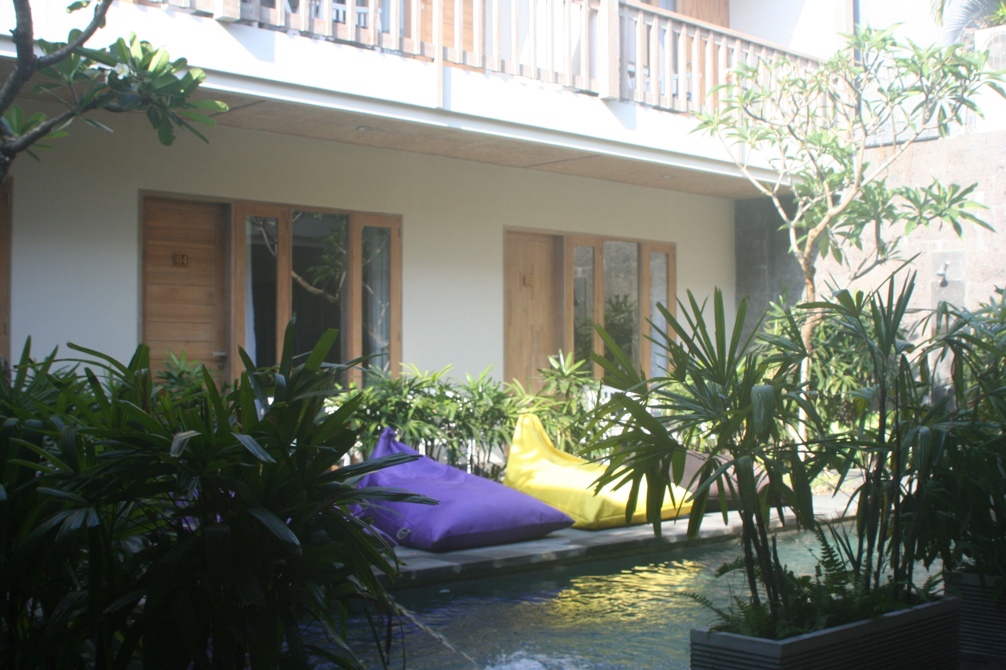 Un área de piscina con un pequeño elemento acuático, rodeada de plantas en macetas y dos coloridos puffs.