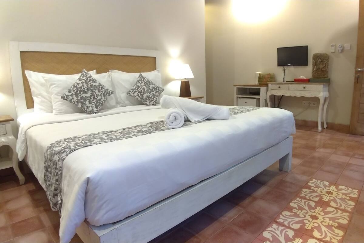 A neatly made bed with a white duvet, grey patterned pillows, and a rolled white towel. There is a small bedside table with a lamp, a desk with a computer and a TV, and a door with a number sign.