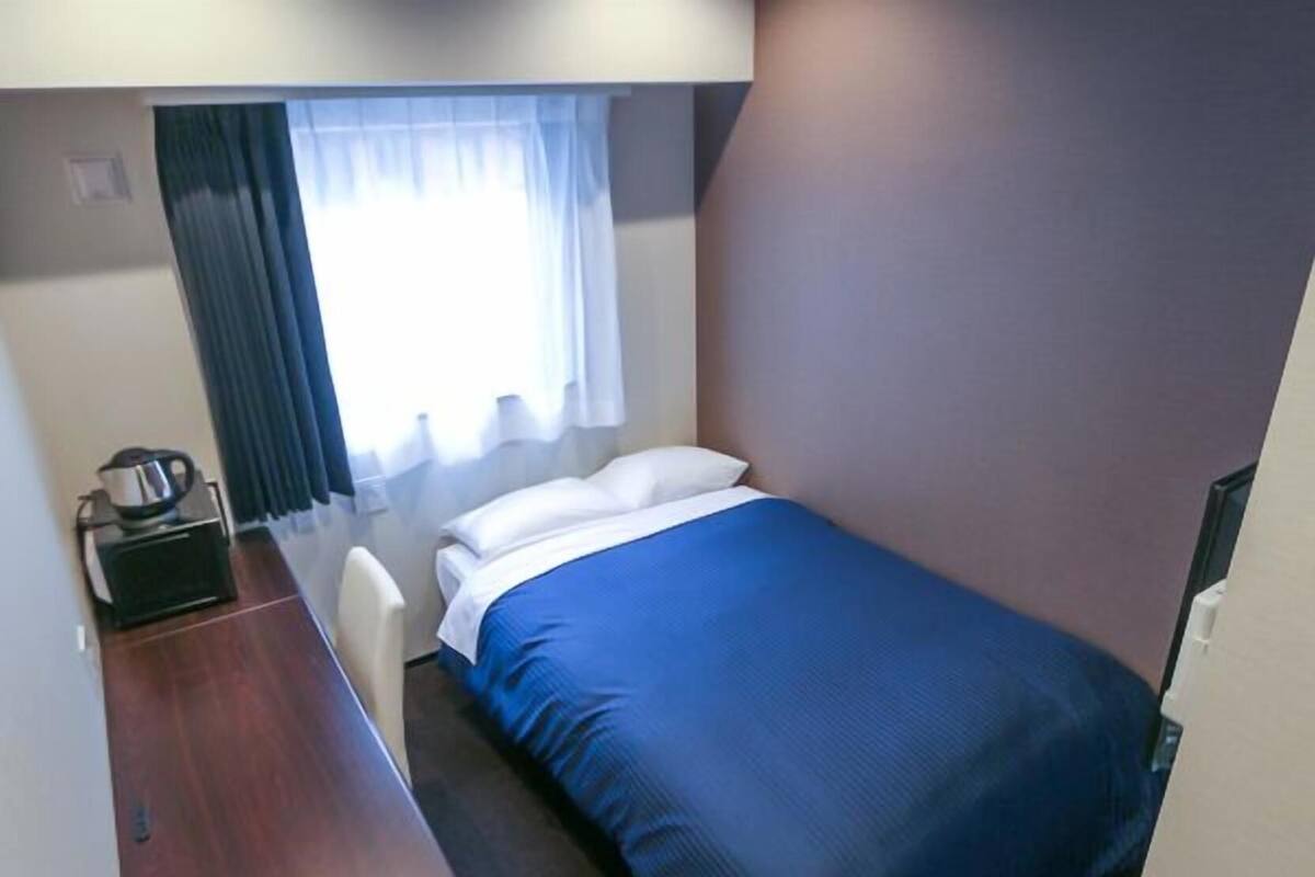 A small, single-bed room with a blue bedspread, a desk with a kettle, and a window with curtains.
