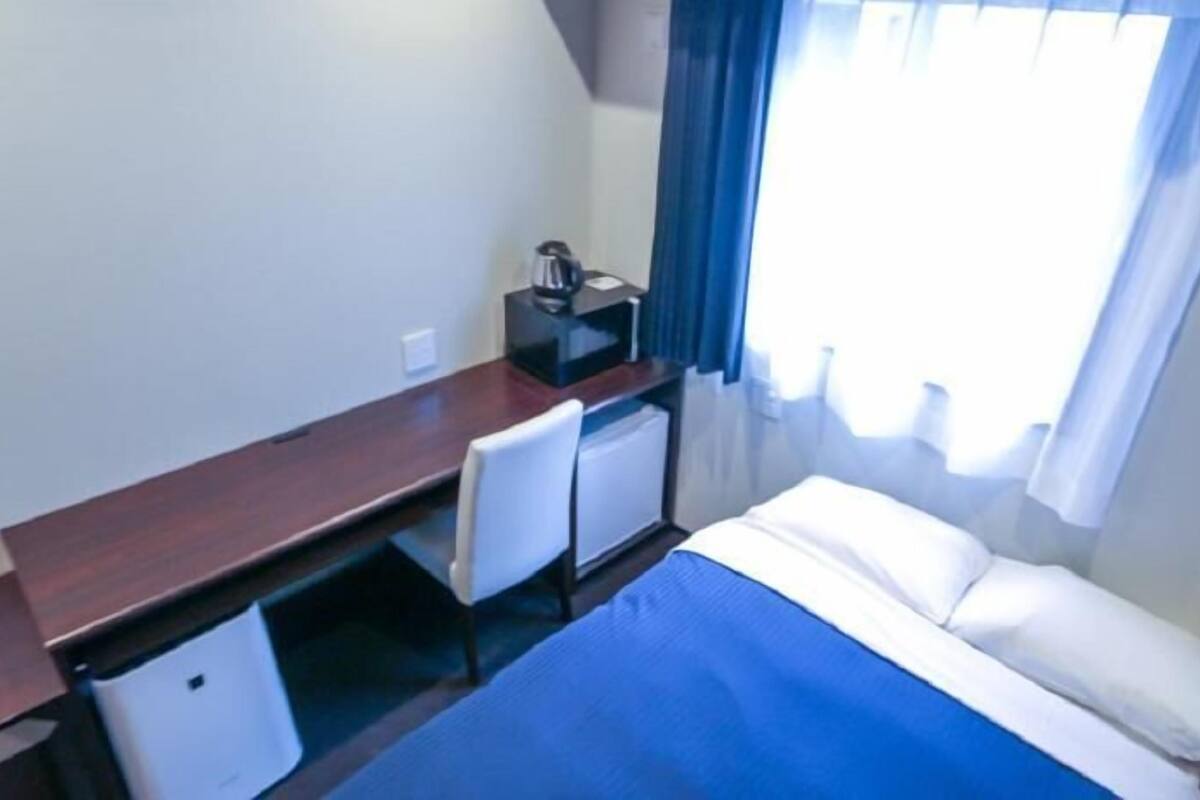 A small, single-room hotel room with a bed, a desk, a chair, a small refrigerator, and a window with curtains.