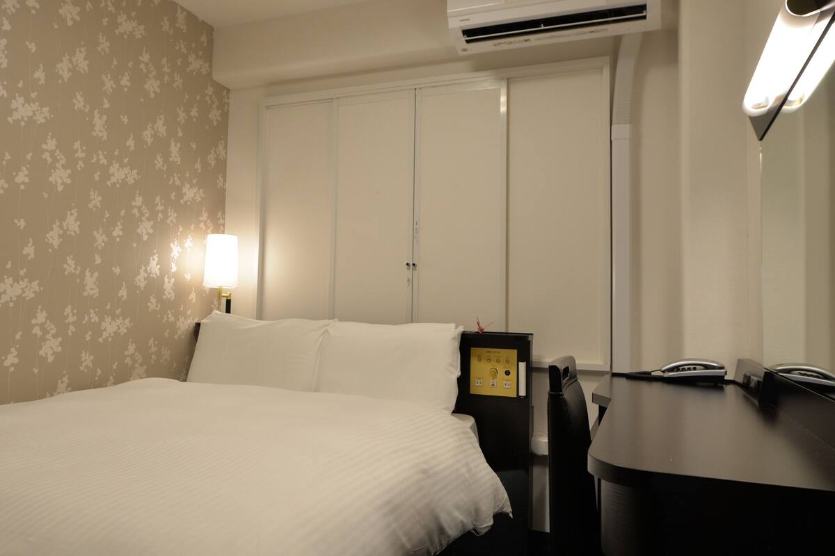 A hotel room with a bed, bedside table, lamp, air conditioning unit, and a desk with a telephone.