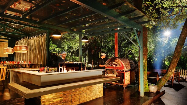 An outdoor restaurant with a stone-tiled floor, a central island with a fire feature, hanging lights, and a rustic wooden oven. - Ayar Jungle Resort (Nainital)
