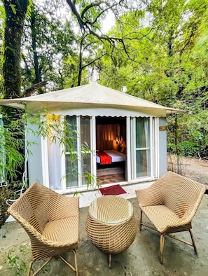 Pequeña construcción de una sola habitación con cama y ventana, rodeada de follaje.