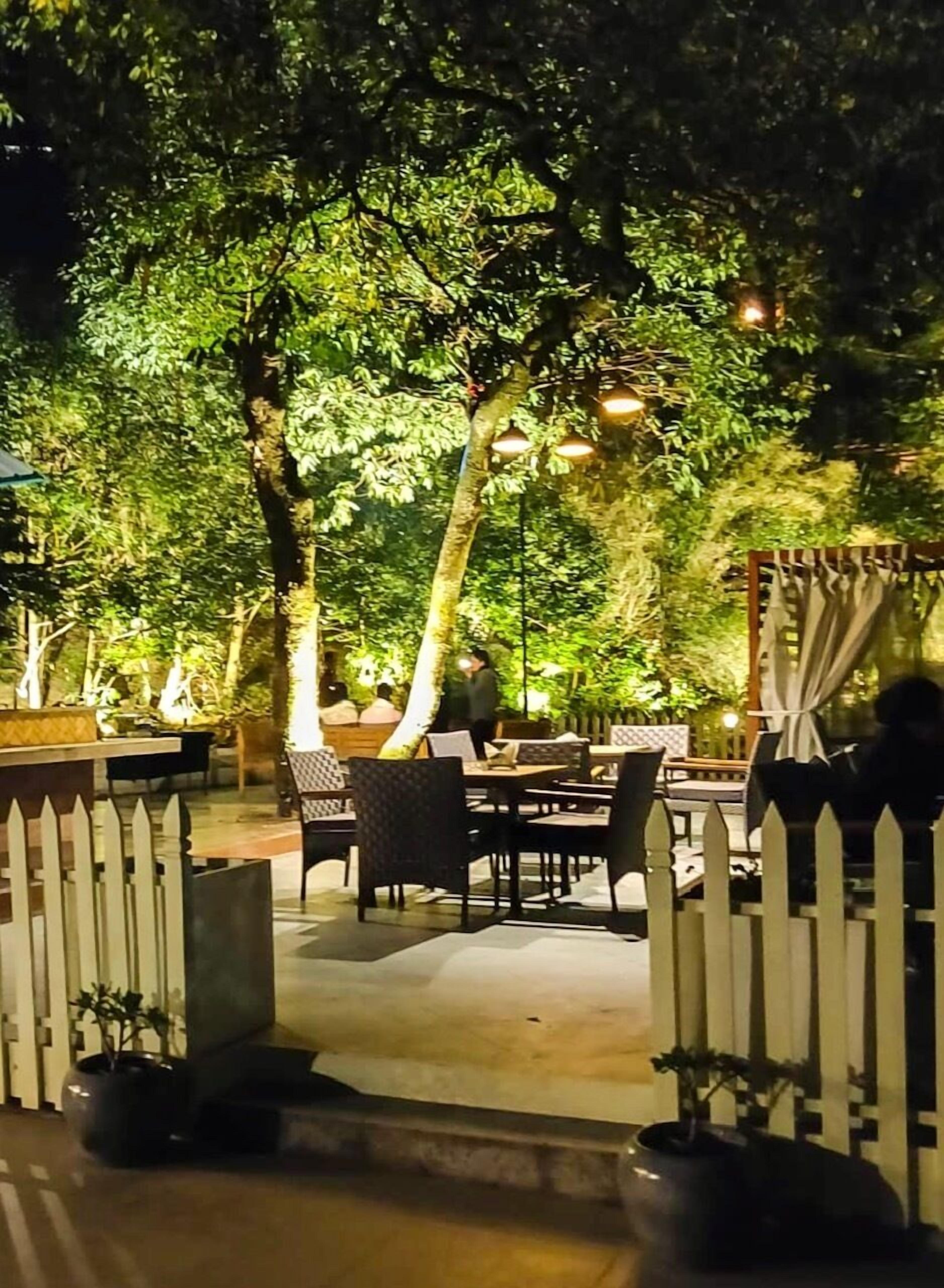 An outdoor dining area with tables and chairs, surrounded by trees and a white picket fence.
