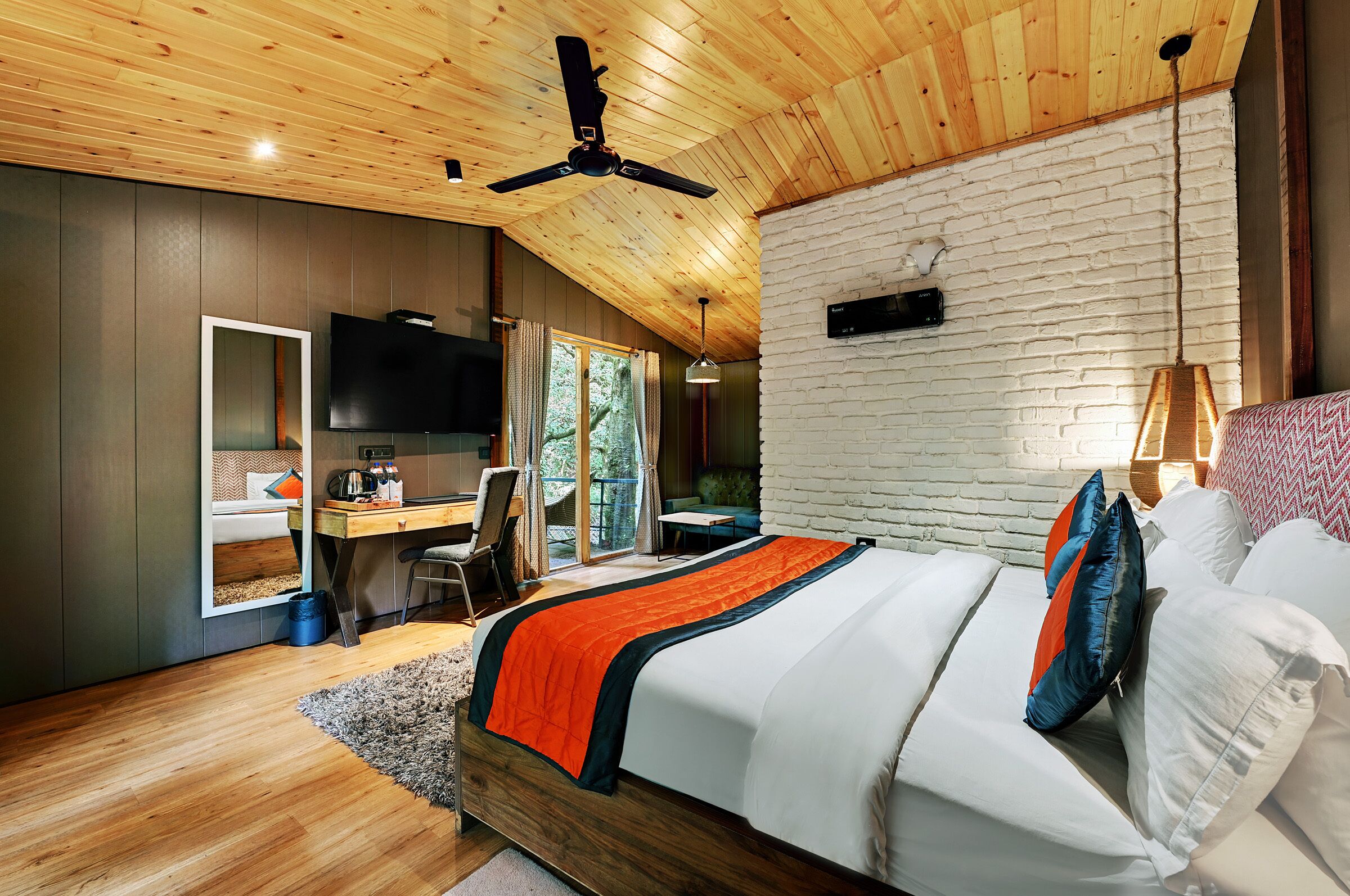 A modern bedroom with a wooden ceiling, a large bed with white and orange bedding, a desk with a TV, and a white brick wall.