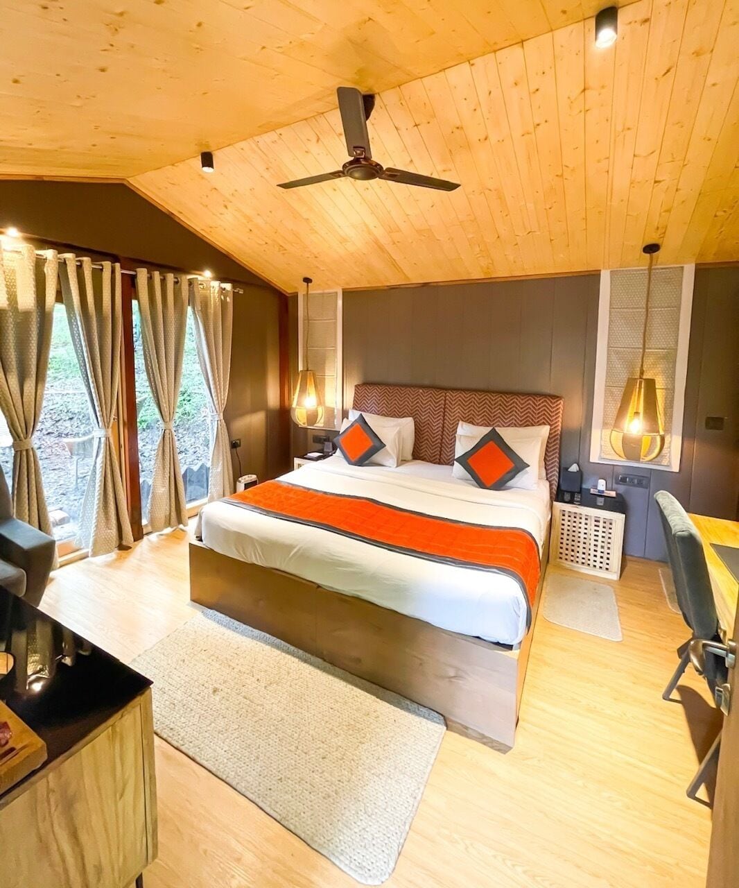 Quarto com teto de madeira, cama com roupa de cama laranja, escrivaninha e uma cadeira.