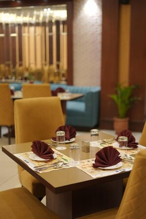 A restaurant setting with tables set for dining, featuring a modern interior design with wooden furniture and a decorative plant. - Hotel Kohinoor Palace (Ludhiana)