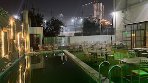 Zona de piscina al aire libre con mesas y sillas, rodeada por un muro bajo e iluminada con luz ambiental.