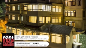 Un hotel de varios pisos con ventanas e balcones iluminados, que muestra la señalización de 'Hotel New Orchid' y 'Voyage Hospitality'.