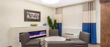 Ein modernes Wohnzimmer mit Kamin, einem runden Tisch, zwei Sesseln, einem gerahmten Bild an der Wand und einem großen Fenster mit Vorhängen.
