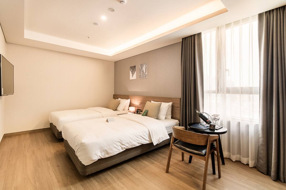 A hotel room with a double bed, a small table with a chair, and a window with curtains.